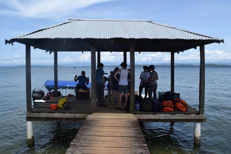 The image shows a wooden pier with a roofed structure over the water. Several people are standing on the pier, some are looking at the camera. There is a boat tied to the pier. The sky is blue and the water is calm.
